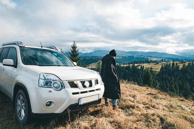 Woman standing near suv car at hill peak looking at beautiful view of mountains road trip