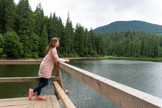 Woman standing on pond and looking at mountain lake with pine forest behind