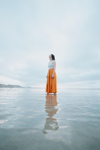 Woman standing on trendy clothes on the beach while reflecting on the water