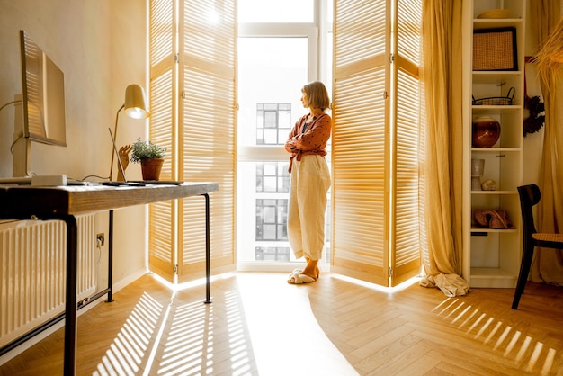 Woman stands alone by the window blinds at home