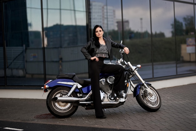 woman stands in front of a motorcycle in front of a glass building in a parking lot near a shopping center.