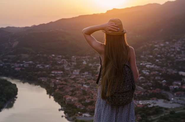 Woman stands on hill looking at city panorama and river at sunset. Back view