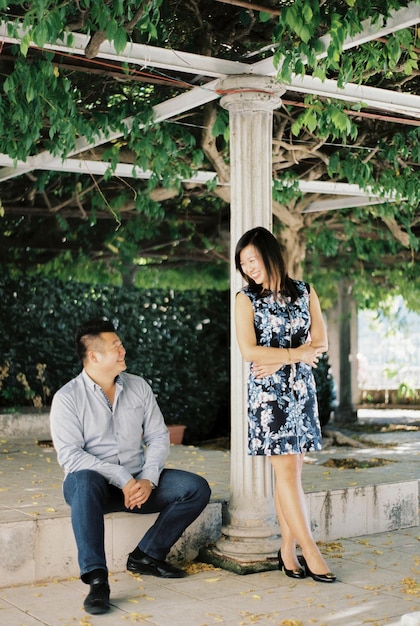 Woman stands leaning on a pergola column next to man sitting on a step