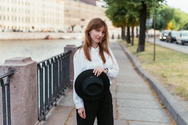 Woman stands near the embankment in the city in the summer holding a hat