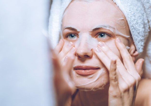 Woman stands near a mirror with a towel on her head and puts on a cosmetic mask