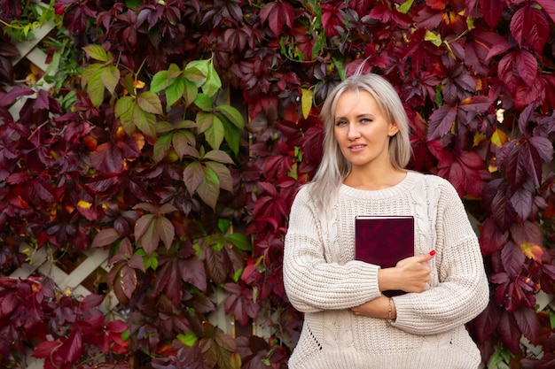 woman stands with book in her hands near vine with red autumn leaves Student holds textbook in hand