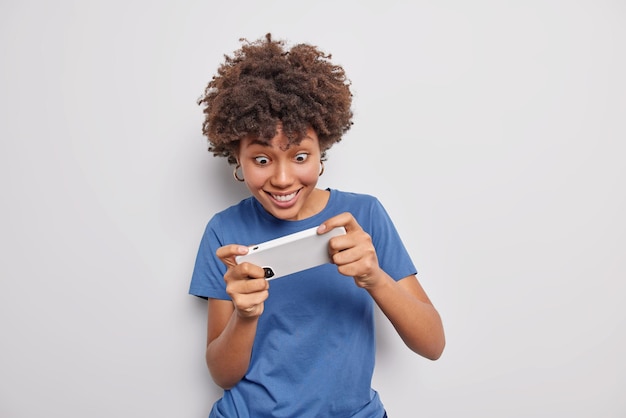 woman stares at smartphone plays games tilts cellular while gaming smiles excited dressed in casual blue t shirt isolated on white. Technology addiction