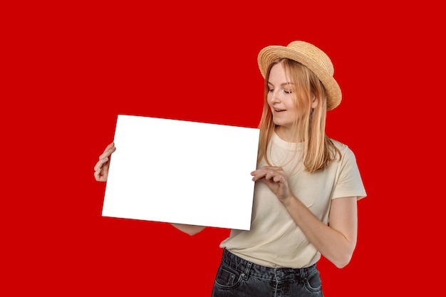 Woman in a straw hat and white tshirt hand holding white blank paper isolated on red studio background with clipping path