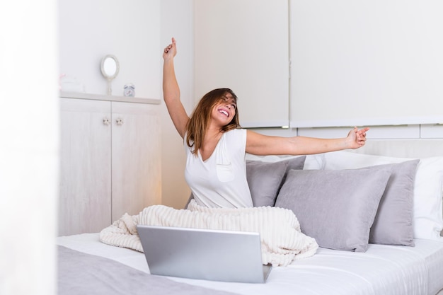 Woman stretching in bed after wake up in morning ready to work from home. Portrait of a young smiling woman sitting with laptop on bed. Freelance working from home concept