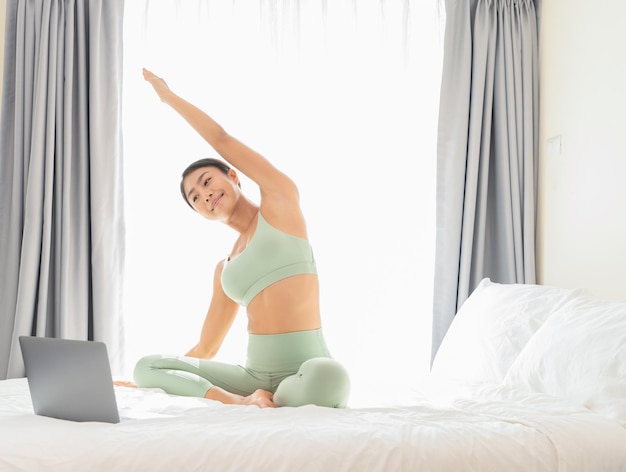 Woman stretching on bed and looking at laptop.Online yoga training at home Healthy and sport concept