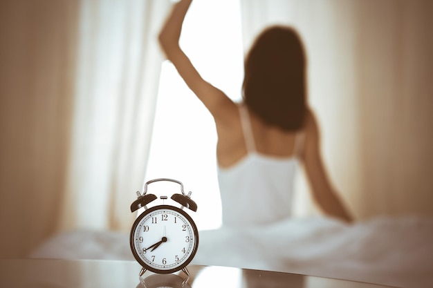 Woman stretching hands in bed after wake up, sun flare . Brunette entering a day happy and relaxed after good night sleep and back view. Concept of a new day and joyful weekend.