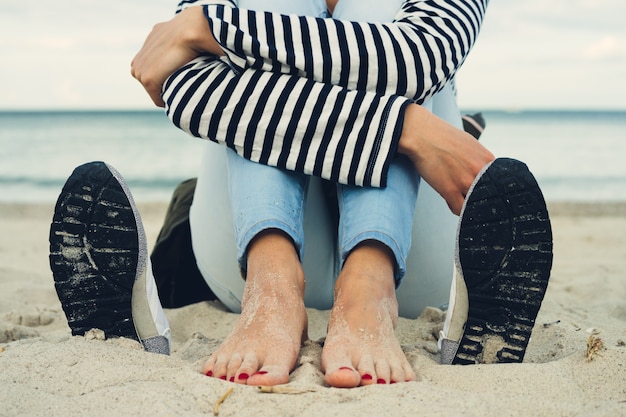 Woman in striped t-shirt and jeans sits barefoot on the beach next to the shoes