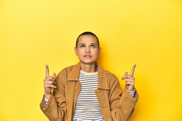 Woman in striped tee and denim shirt yellow backdrop pointing upside with opened mouth