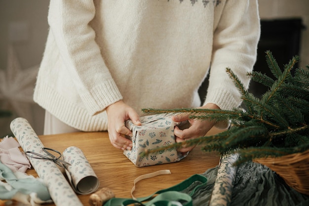 Photo woman in stylish sweater wrapping christmas gift in paper on wooden table with festive decorations in decorated scandinavian room merry christmas hands packing present close up