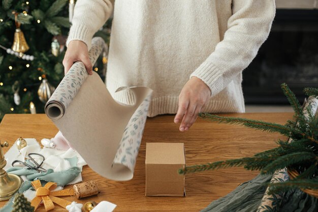 Photo woman in stylish sweater wrapping christmas gift in paper on wooden table with festive decorations in decorated scandinavian room merry christmas hands packing present close up