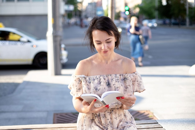 A woman in a summer dress sits on a bench in the middle of the street and reads a book