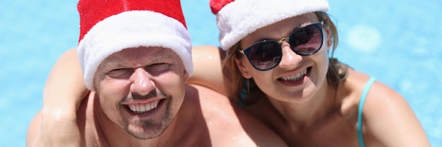 Woman in sunglasses and santa claus hat hugging man on side of swimming pool