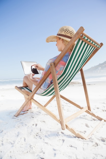 Woman in sunhat sitting on beach in deck chair using tablet pc