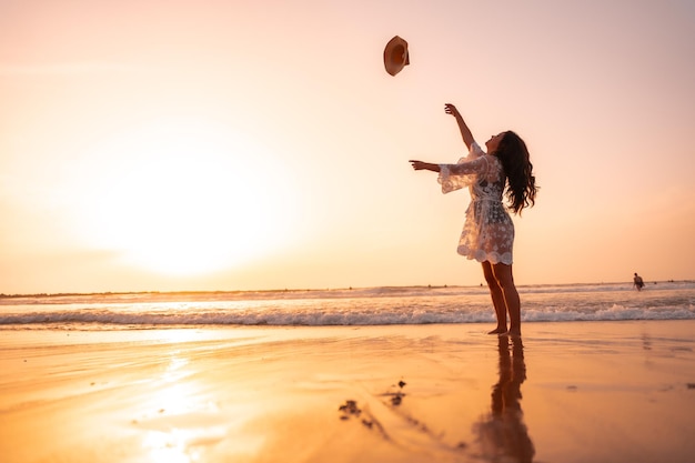 A woman in the sunset in a white dress at the sea in summer throwing the hat on vacation