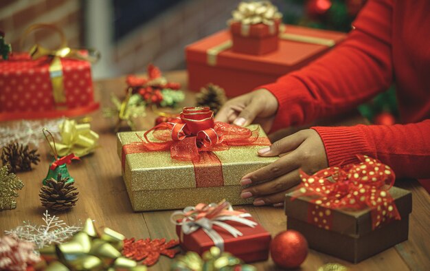 Woman on sweater put golden luxurious present box with gorgeous bow on table of beautiful ornament and colorful ribbon to prepare wonderful gift for home party decoration of Christmas holiday festival