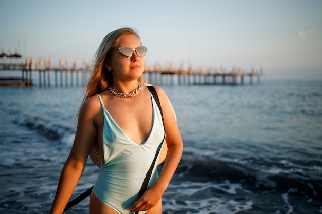 A woman in a swimsuit and a hat with glasses walks along the beach at sunset. The concept of sea recreation. Selective focus