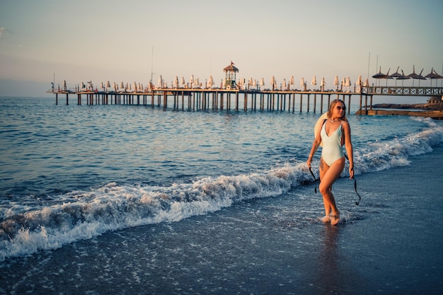 A woman in a swimsuit and a hat with glasses walks along the beach at sunset The concept of sea recreation Selective focus