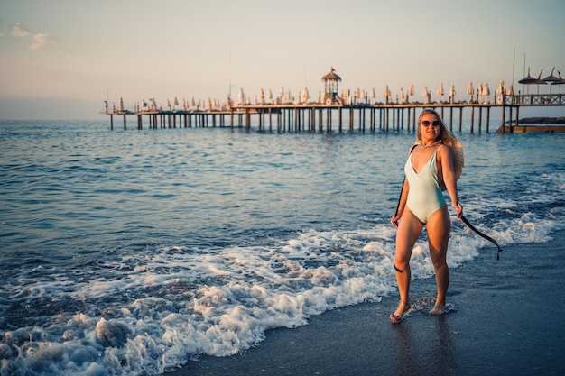 A woman in a swimsuit and a hat with glasses walks along the beach at sunset The concept of sea recreation Selective focus