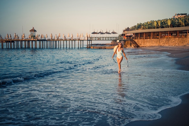 A woman in a swimsuit and a hat with glasses walks along the beach at sunset The concept of sea recreation Selective focus