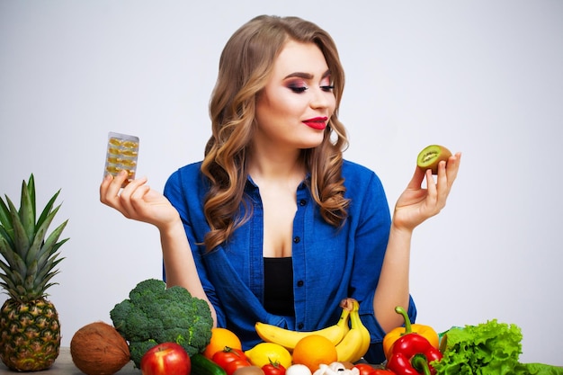 Woman at a table holding a kiwi and pills on a background of fruit and vegetables.