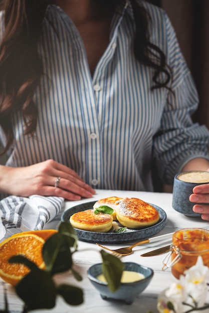 Woman at the table with a beautiful and tasty breakfast. Cottage cheese pancakes on a plate, cup with coffee and oranges on a white wooden background.