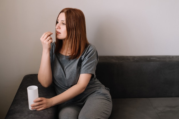 Woman takes a vitamin pill sitting on a couch in the living room at home. Coronavirus problem and covid-19 protection concept.