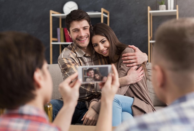 Woman taking photo of loving couple on mobile phone at home