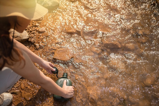 Woman taking pure water to bottle from mountain stream during trekking in mountains Hiker crouching on rocks filling bottle up with cold mountain water