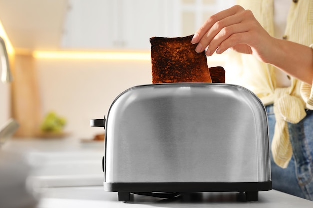 Woman taking slice of burnt bread from toaster in kitchen closeup