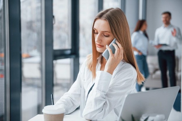 Woman talking by phone in front of group of young successful team that working and communicating together indoors in office