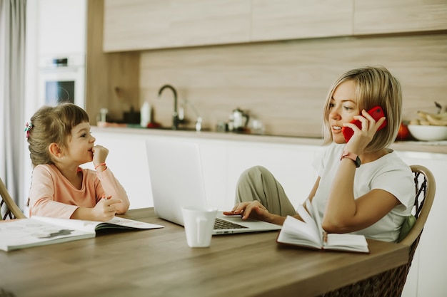 Woman talking by phone while working at home and her little daughter sitting next to her