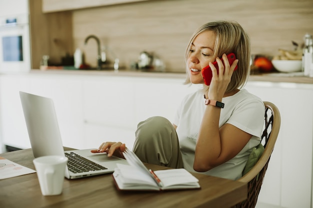 Woman talking by phone while working at home Shes sitting in the kitchen using laptop