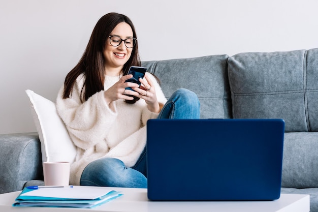 Woman talking with mobile phone while working with laptop sitting on a couch at home