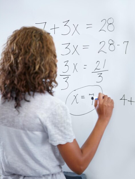 Photo woman teacher and writing on whiteboard for mathematics numbers or equations in classroom rear view of female person educator or lecturer with sum on board for learning or problem solving class