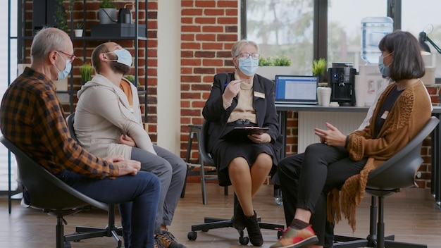 Woman therapist giving advice to people at aa group meeting, wearing face mask. Psychiatrist having conversation with patients about alcoholism and recovery during coronavirus pandemic.