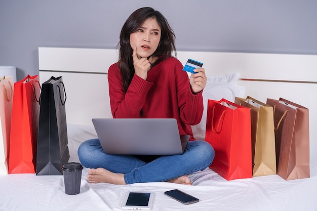 Woman thinking and using laptop computer for online shopping with credit card on bed