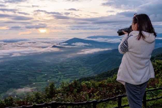 Woman tourist are using a DSLR camera photographing nature landscape the sun fog mountain in the winter during sunrise on high viewpoint at Phu Ruea National Park, Loei province, Thailand