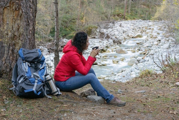 Woman tourist drinks coffee on a halt in the woods by the river.