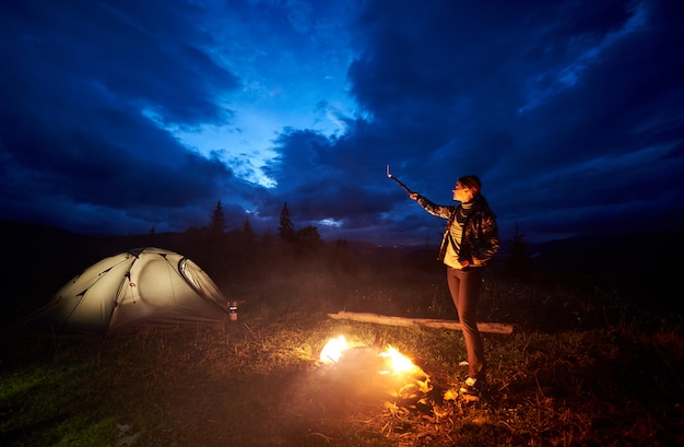 Woman tourist having a rest at night camping in the mountains, standing near burning campfire and illuminated tourist tent, pointing at evening cloudy sky