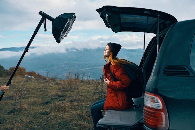 Woman tourist with a backpack sits on the trunk of a car in the mountains in autumn in nature