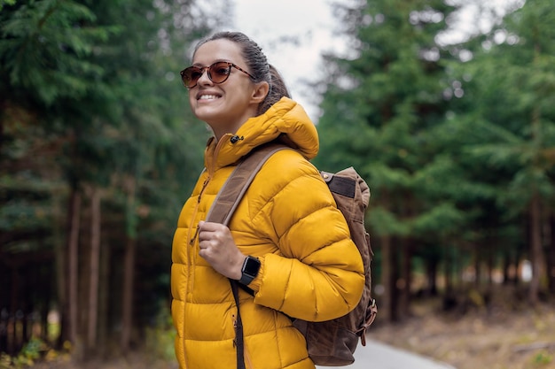 Woman tourist in yellow down jacket with leather retro tourism backpack have a walk in nature backgr