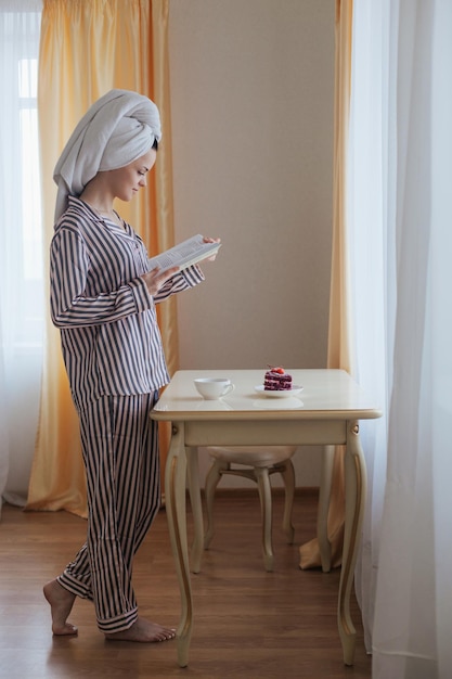 Woman in towel stands near the table read interesting book drink tea with cake in the morning