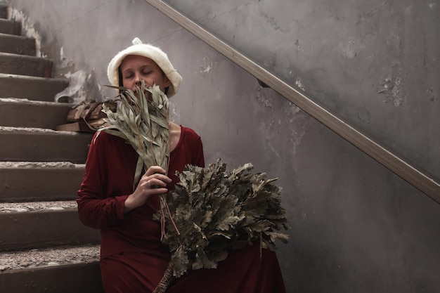 Woman in traditional dress with bath brooms in traditional steam sauna banya on stairs outdoors in spa complex Russian bathhouse