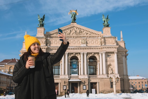 Woman traveler drinking coffee to go taking selfie in front of opera building lviv city ukraine