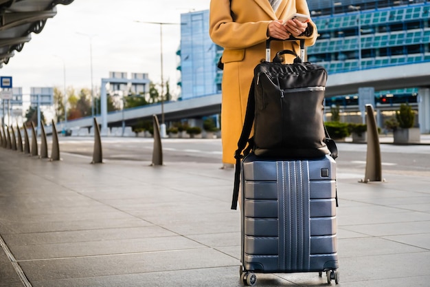 Woman traveler in long yellow coat with handbag and suitcase orders taxi cab standing near internati...